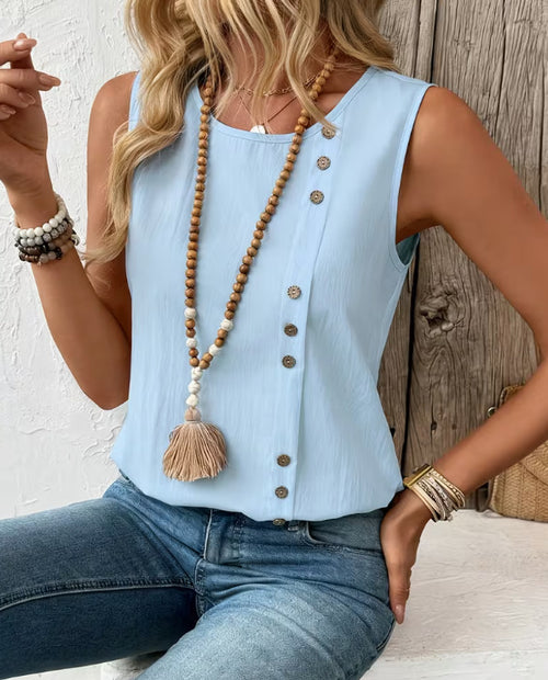 Woman wearing a light blue sleeveless top with buttons and jeans, sitting against a wooden wall.