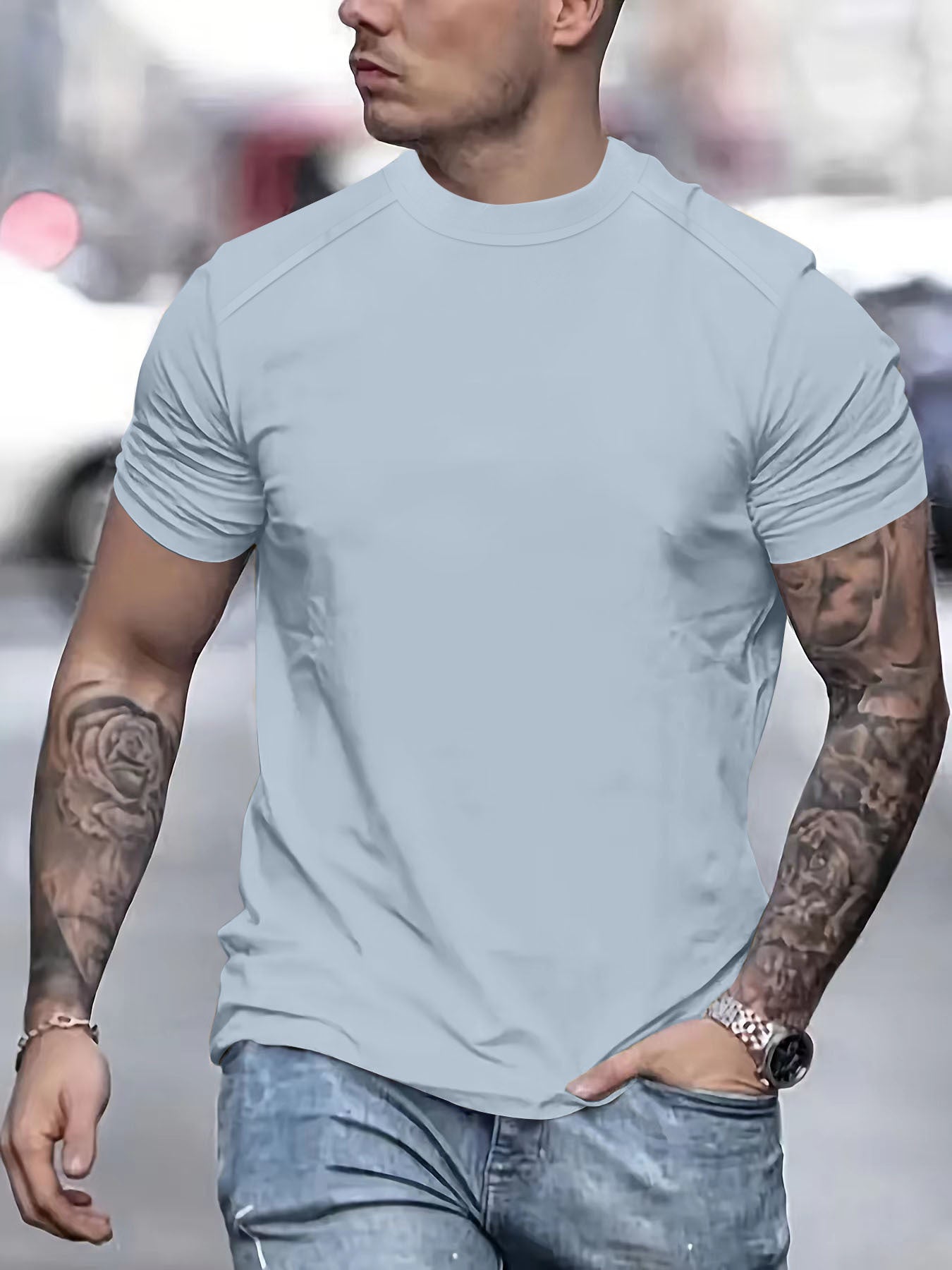 Man wearing a light blue t-shirt and jeans with tattoos on arms, blurred background