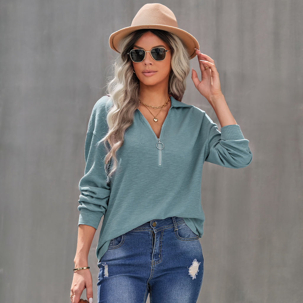 Woman wearing a teal sweater, beige hat, and sunglasses against a gray background