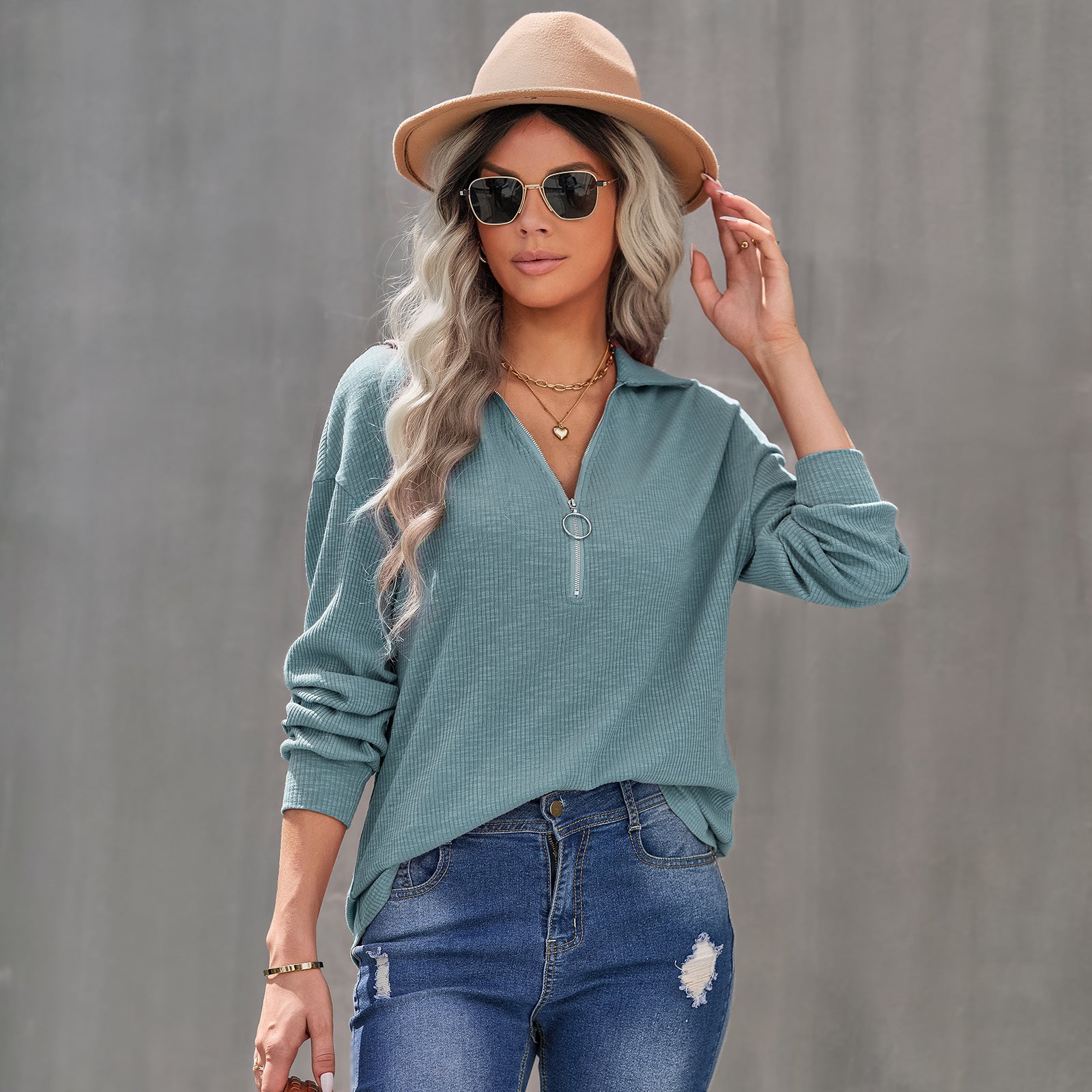 Woman wearing a teal sweater, beige hat, and sunglasses against a gray background