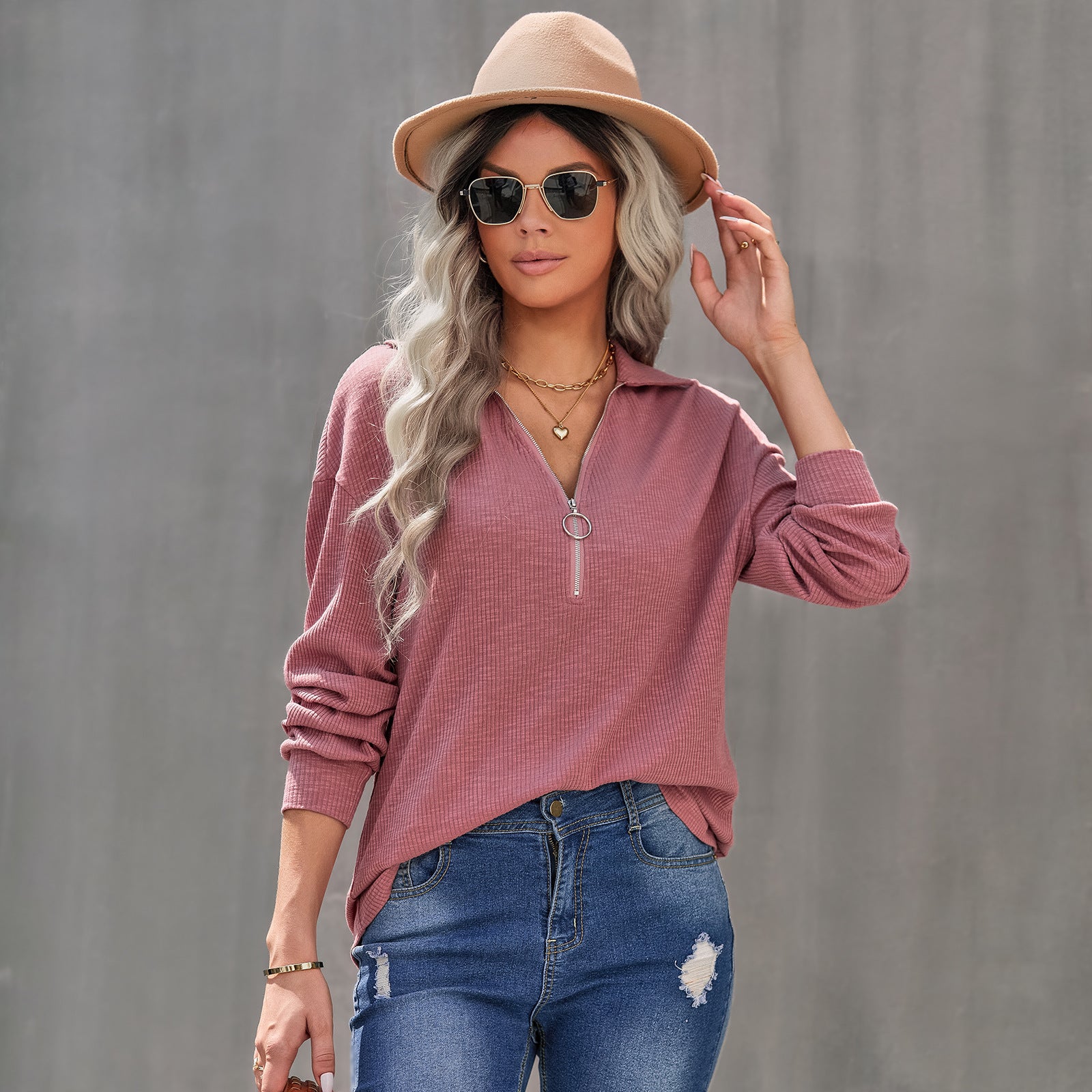 Woman wearing a pink long-sleeve top, blue jeans, and a beige hat against a gray background